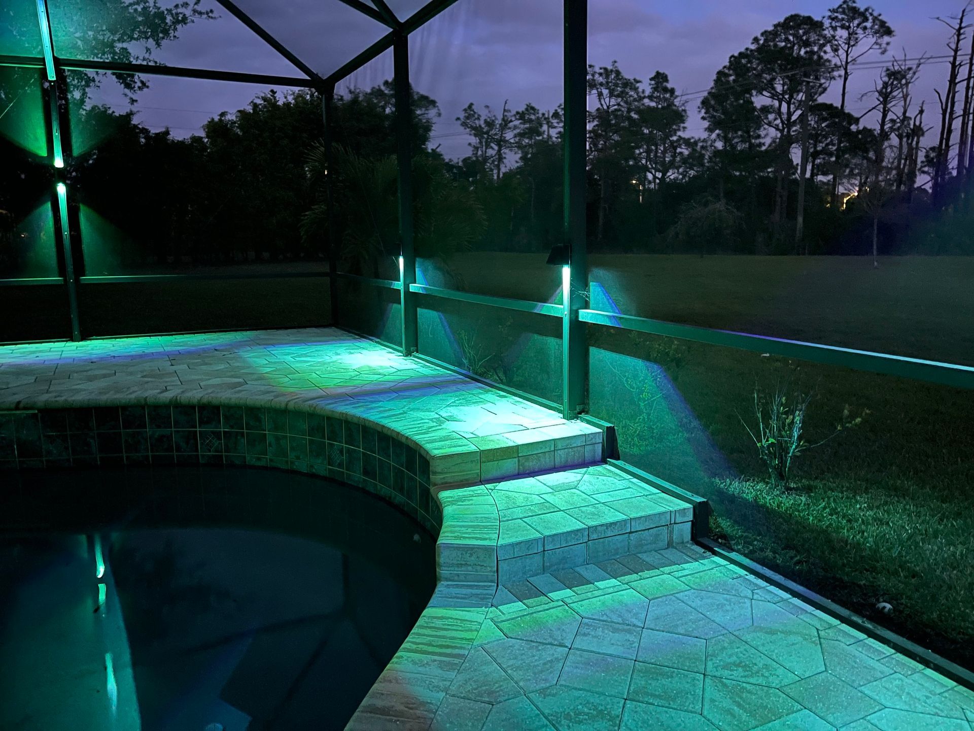 Pool area at night illuminated with green lights along the walls and steps, with trees in the background.
