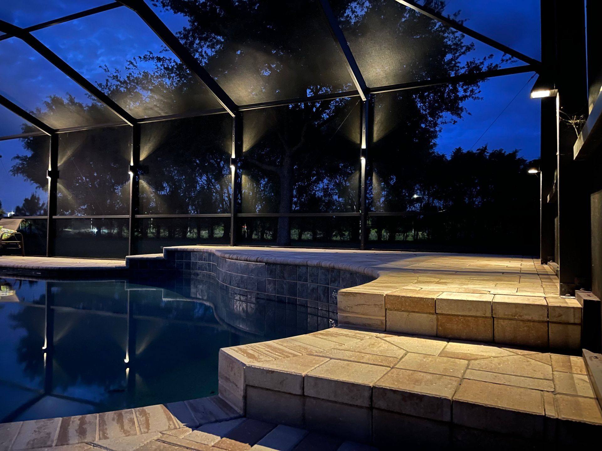 Nighttime view of a pool with lighted enclosure. Steps, tiled deck, and blue sky visible.