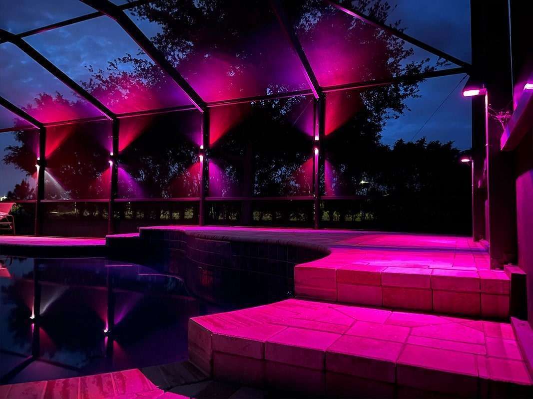 Nighttime pool area with pink and purple lighting illuminating the pool, patio, and overhead trellis.