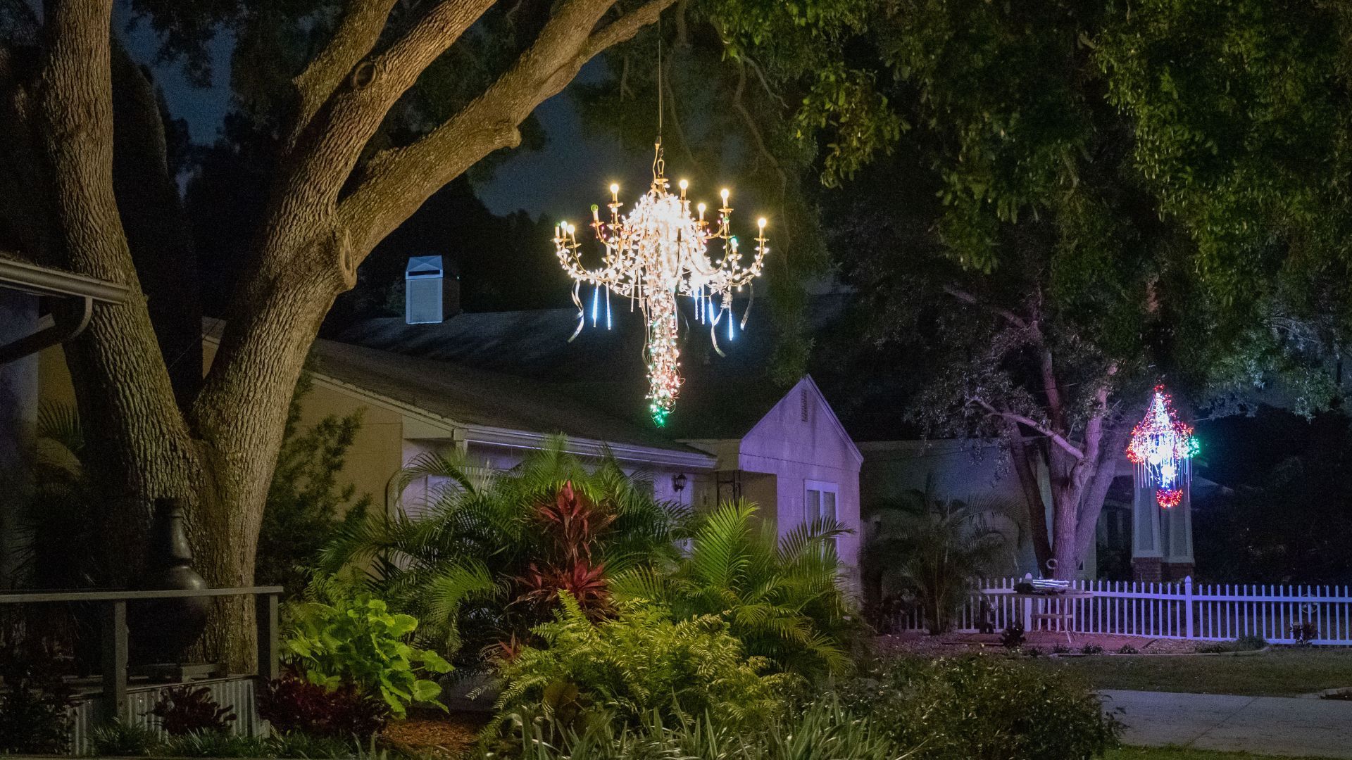 A lit-up chandelier decoration hangs from a tree in front of a house at night.