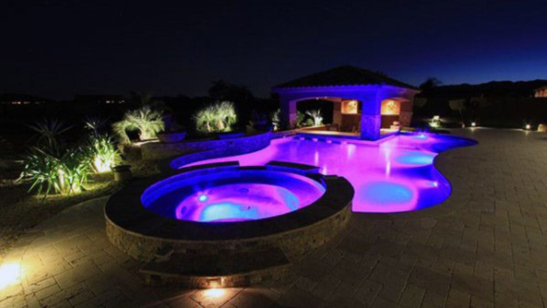 Night view of a luxury pool and hot tub lit with blue and purple lights, next to an open-air cabana.