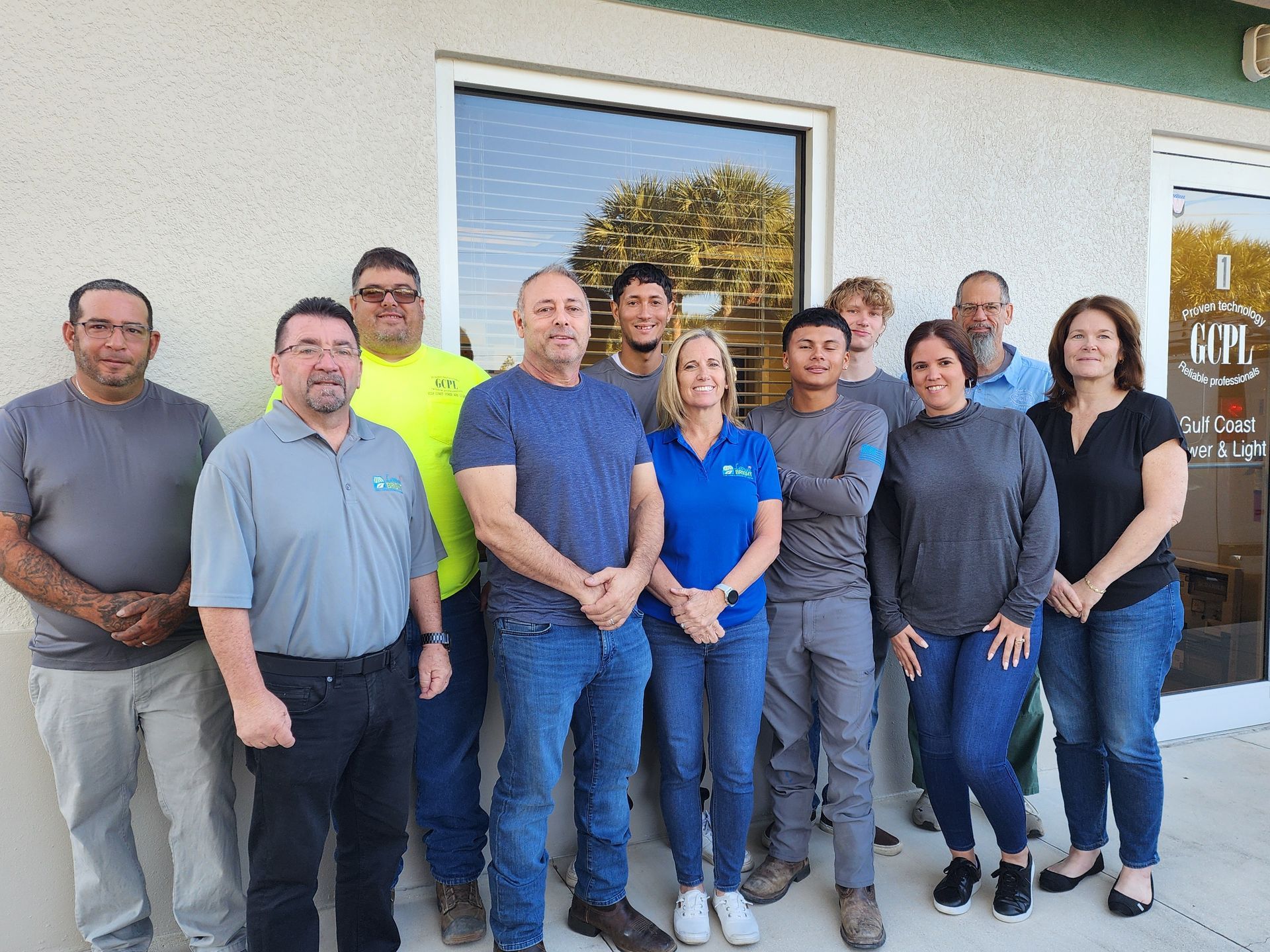 Group of people standing outside a building with a window and sign. Most are smiling, wearing casual clothes.