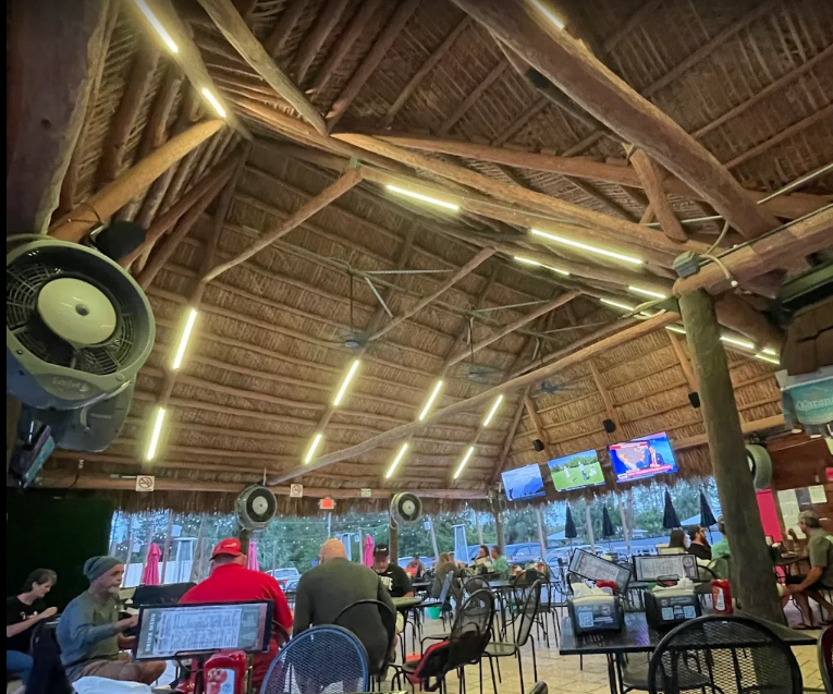 A thatched-roof outdoor bar with people seated at tables, watching TVs.