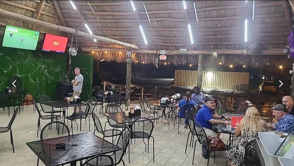 Outdoor restaurant with tables, chairs, and patrons. TVs are mounted on a green wall.
