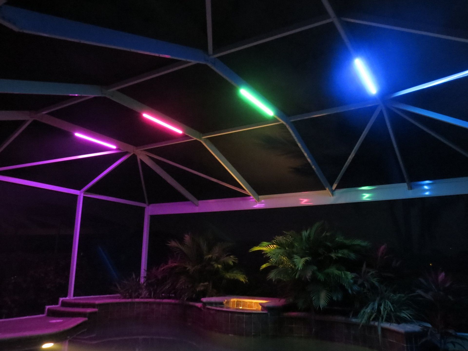 Pool enclosure lit with purple, green, and blue lights, with palm trees and a dark sky.