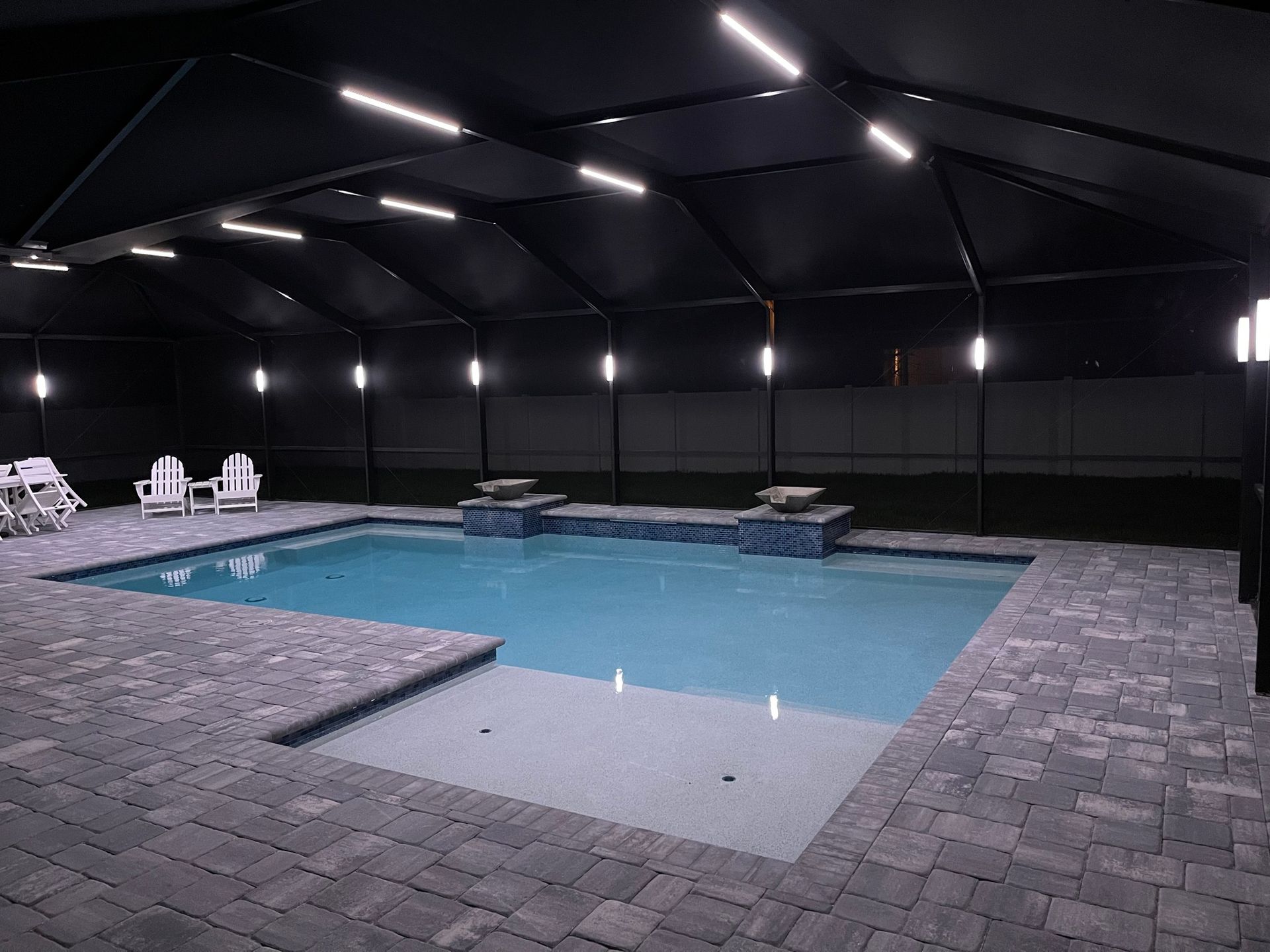 Indoor pool with dark roof and lighting. Blue water surrounded by gray pavers.