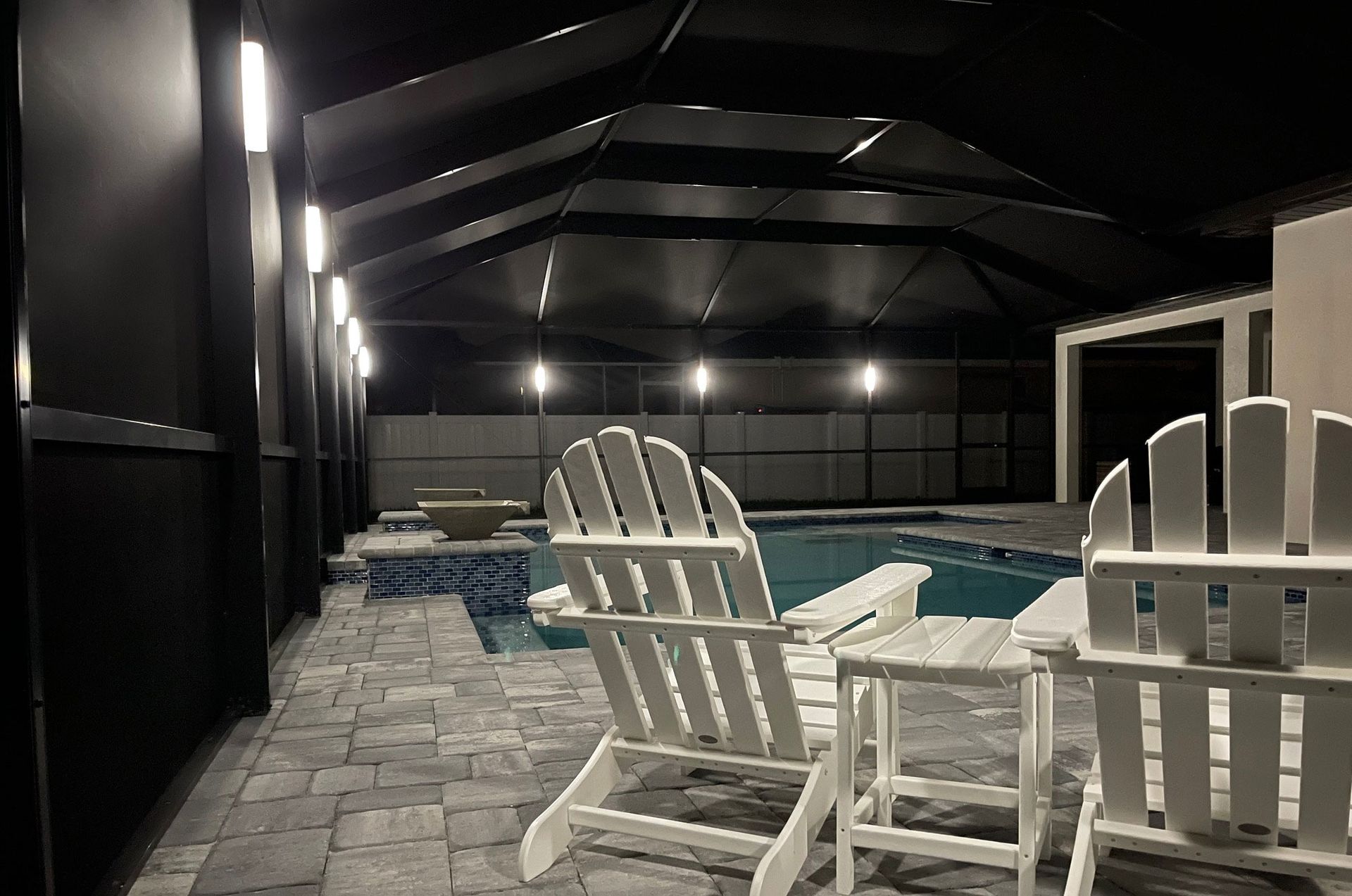 Two white chairs beside a pool under a black screened enclosure, lit by overhead lights.