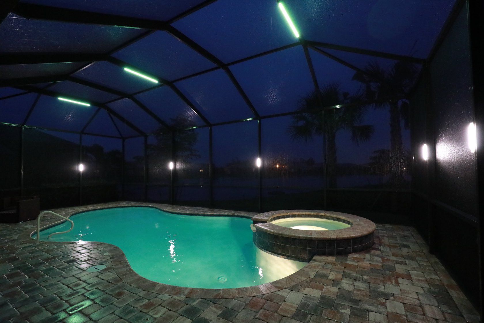 Swimming pool and hot tub under a screened enclosure at dusk, illuminated with green and white lights.