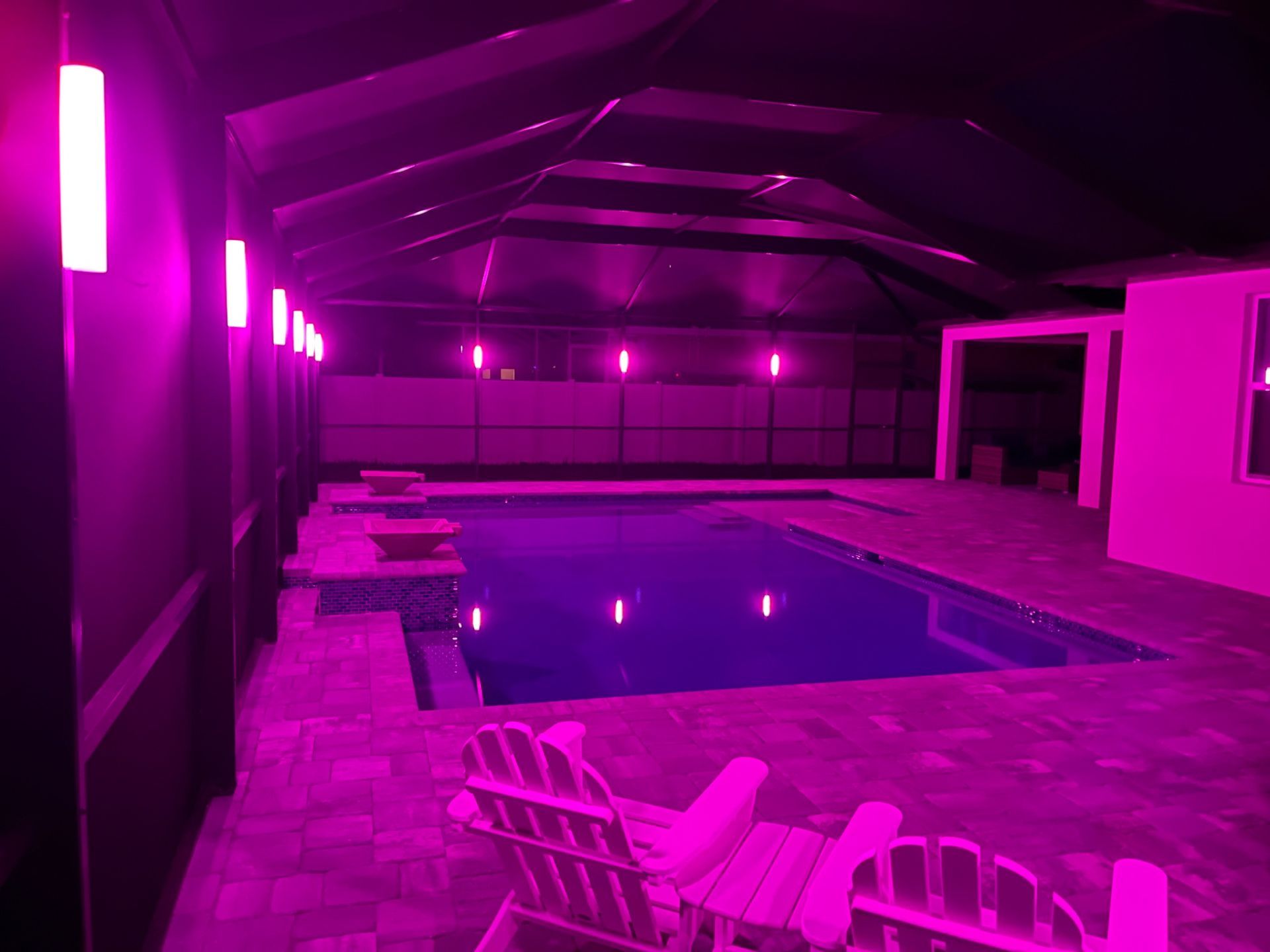 A pink-lit indoor pool area with a rectangular pool, chairs, and wall lights.