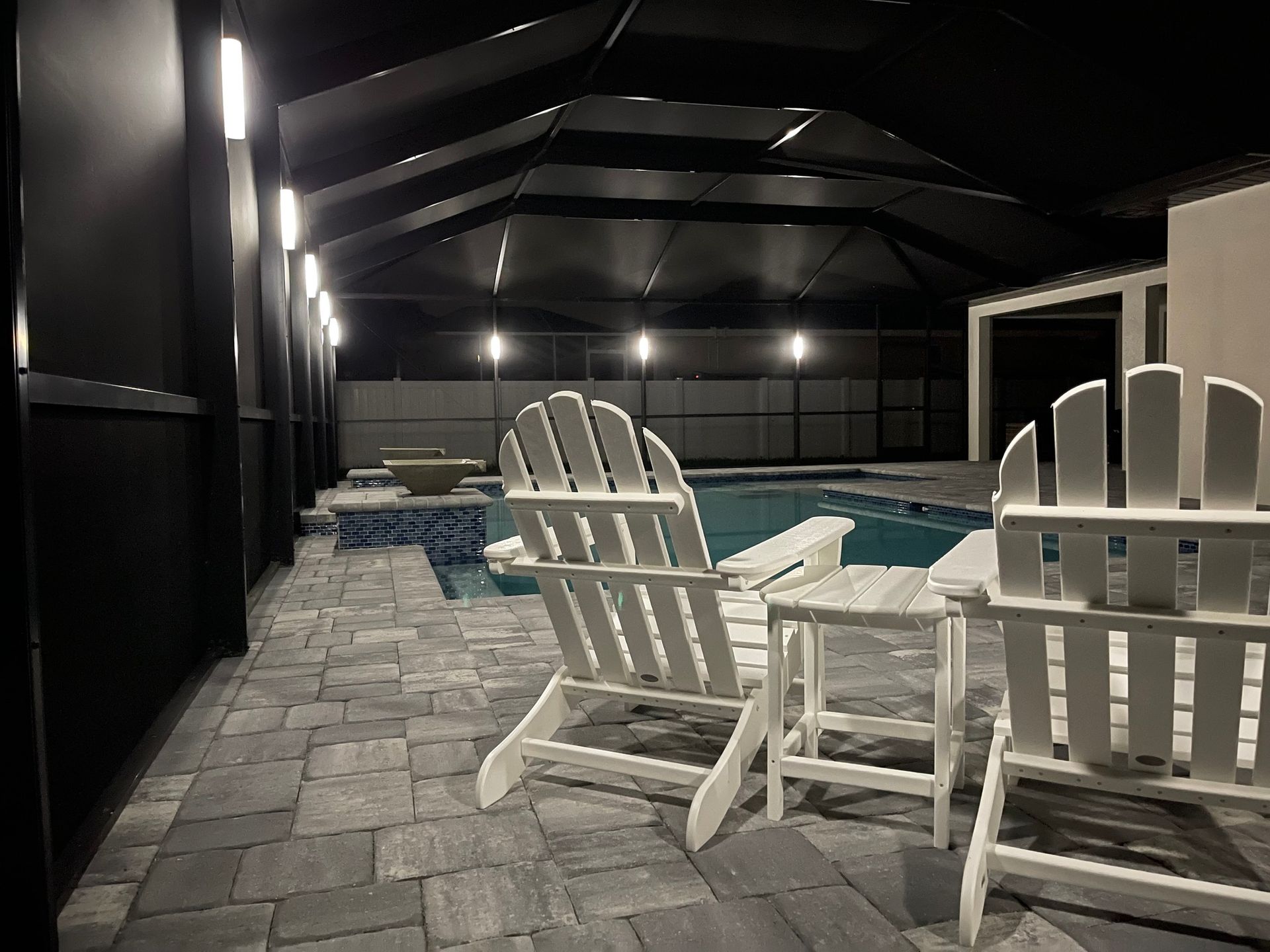 Patio with white chairs, small table, pool, and overhead shade structure at night.