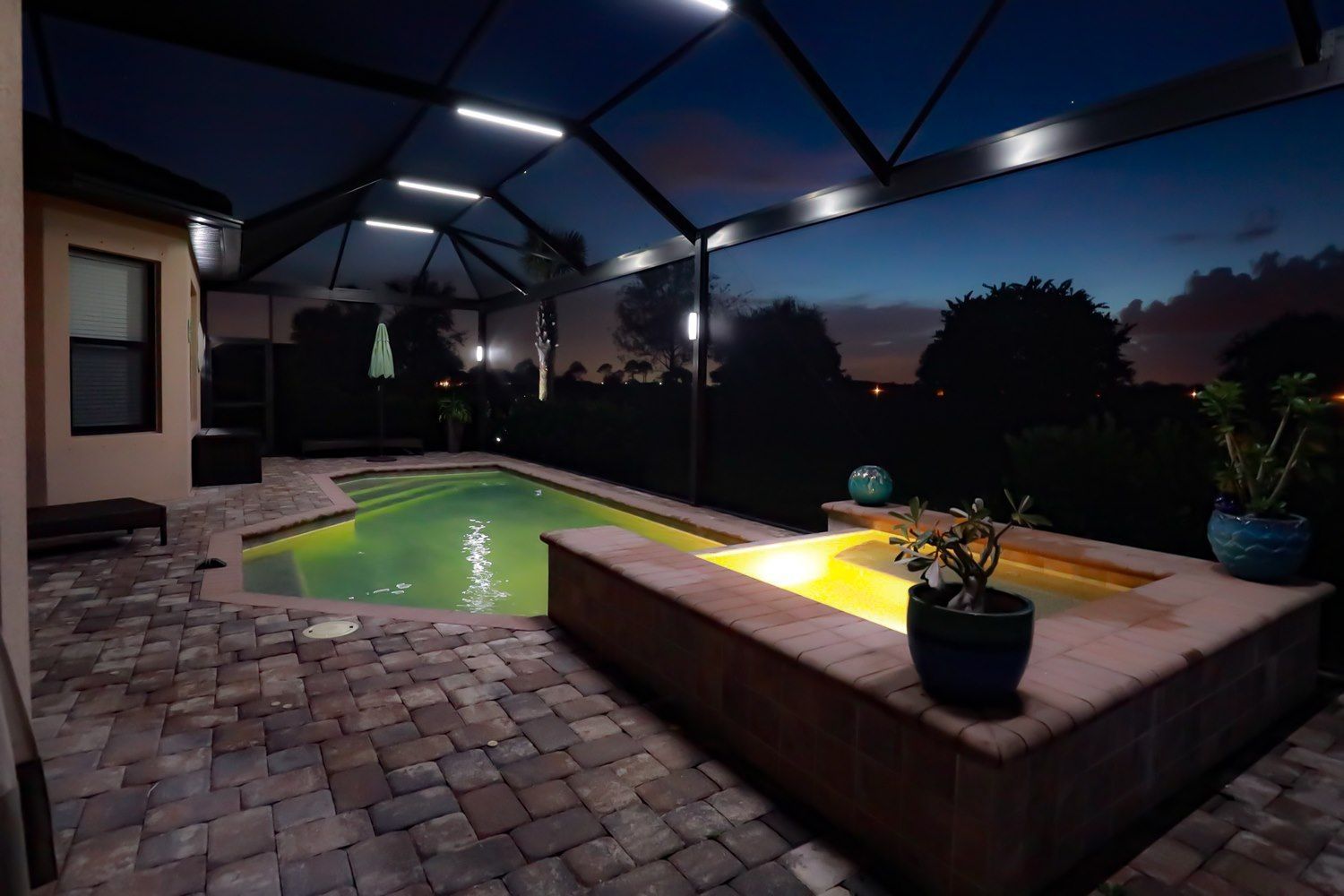 Nighttime view of a backyard pool and spa with overhead lighting, a brick patio, and a dark sky.