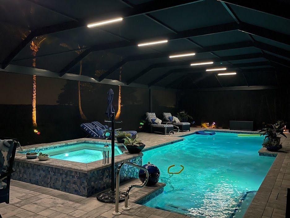 Indoor pool with hot tub, lounge chairs, and overhead lighting. Teal water and dark surroundings.