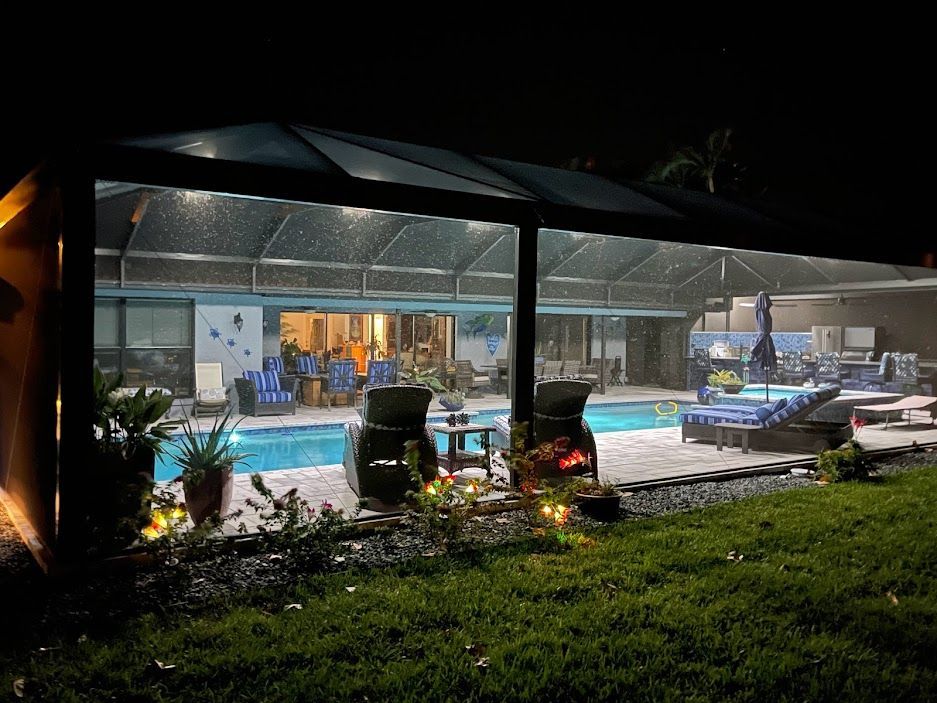 Night view of a pool area with a covered patio lit by overhead lights. The house is visible in the background.