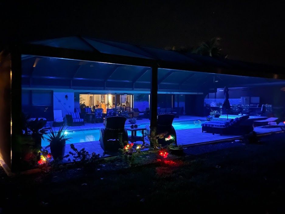 Nighttime backyard pool scene with blue lighting under a patio cover, illuminating the pool and surrounding area.