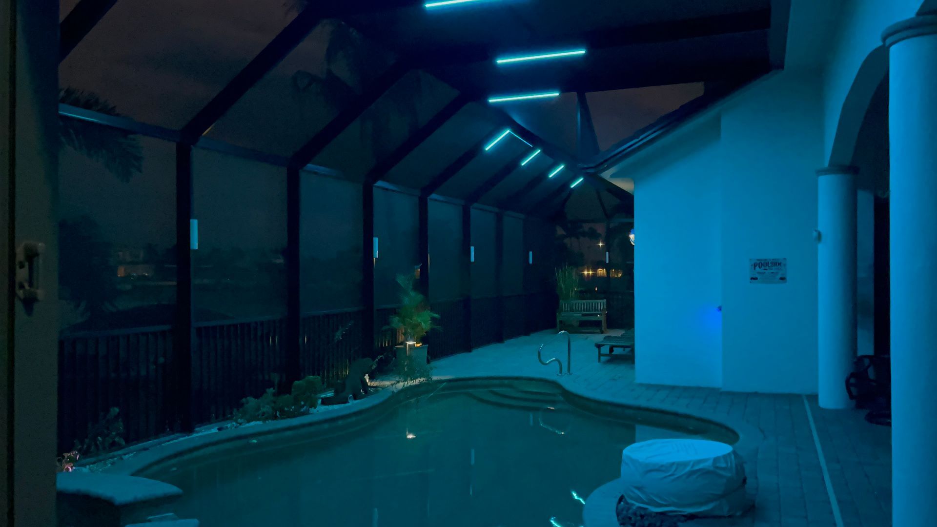 Pool area at night illuminated with blue lights. Pool, screened enclosure, and house are visible.
