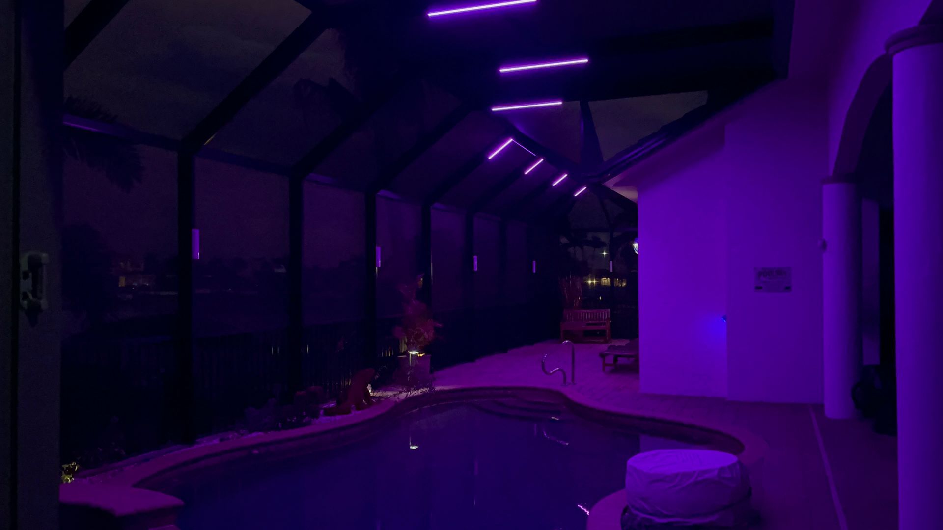 Purple-lit poolside at night. Reflective water, white building, and glass roof with light fixtures.