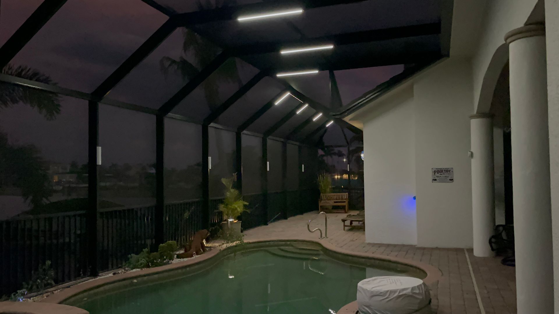 Night view of a screened-in pool area with overhead lighting. The pool is surrounded by a patio and a white stucco building.