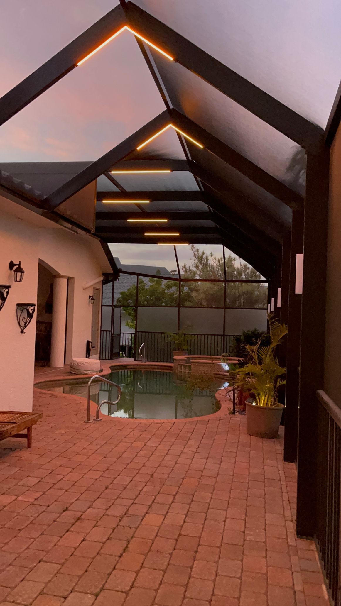 Brick patio leads to a pool under a covered structure with warm lighting. Trees and sky visible beyond.