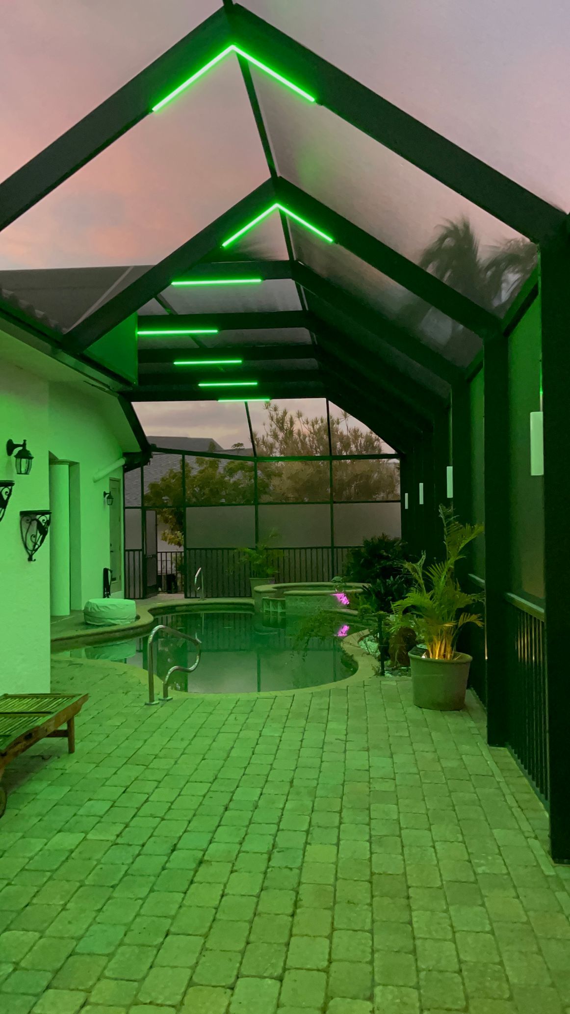 Green-lit pool area with a covered walkway. Pink and orange sky, plants, and pool.