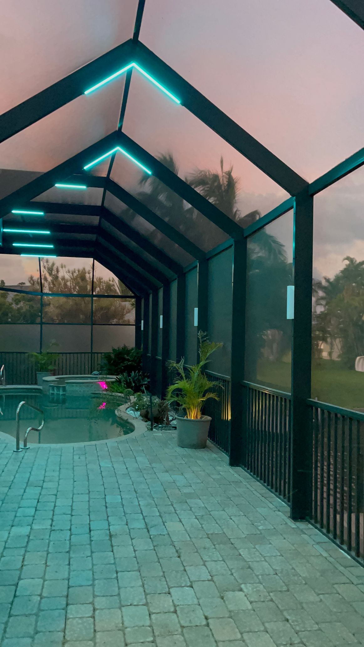 Screened-in pool patio with teal-lit roof and brick floor at dusk. Plants and pool in view.