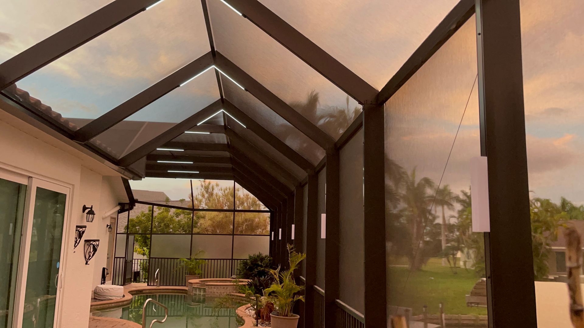 Screened patio with clear roof and string lights overlooking a pool and yard at dusk.