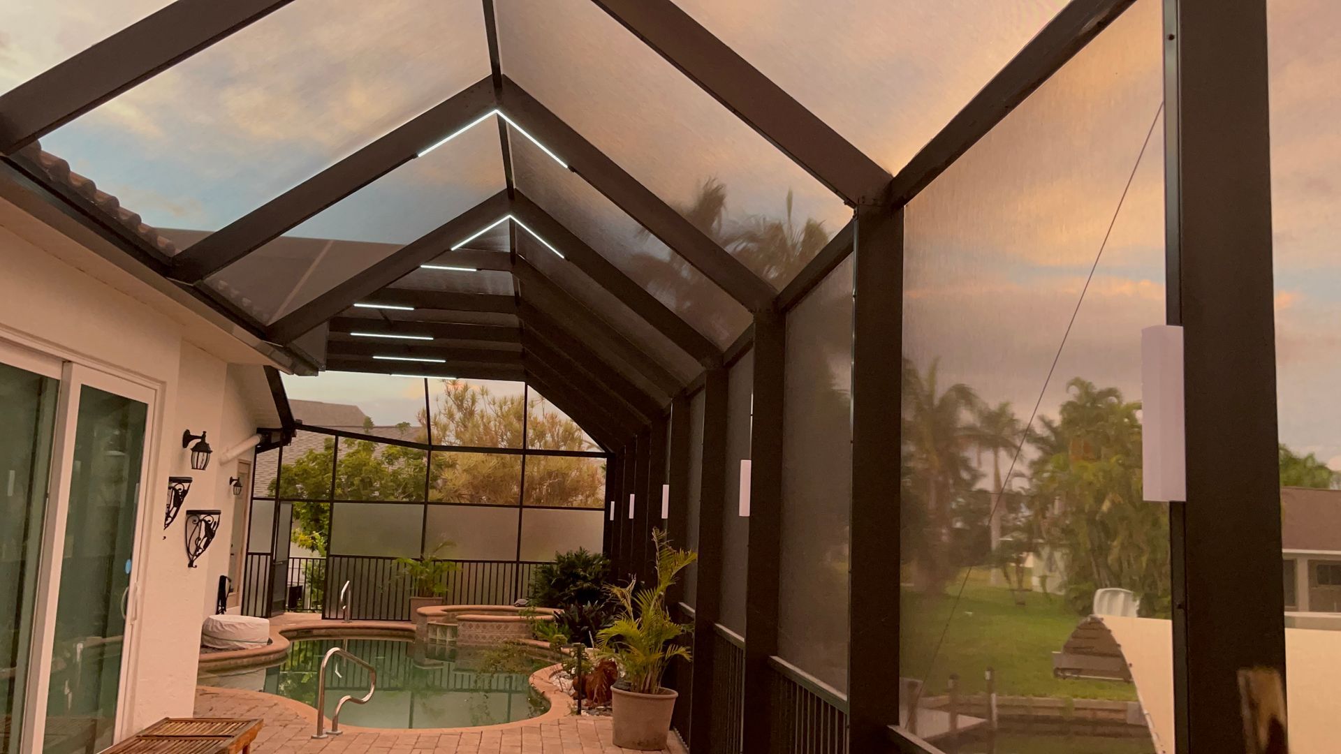 A screened patio with a pool in the background, under an angled glass roof, with an overcast sky.