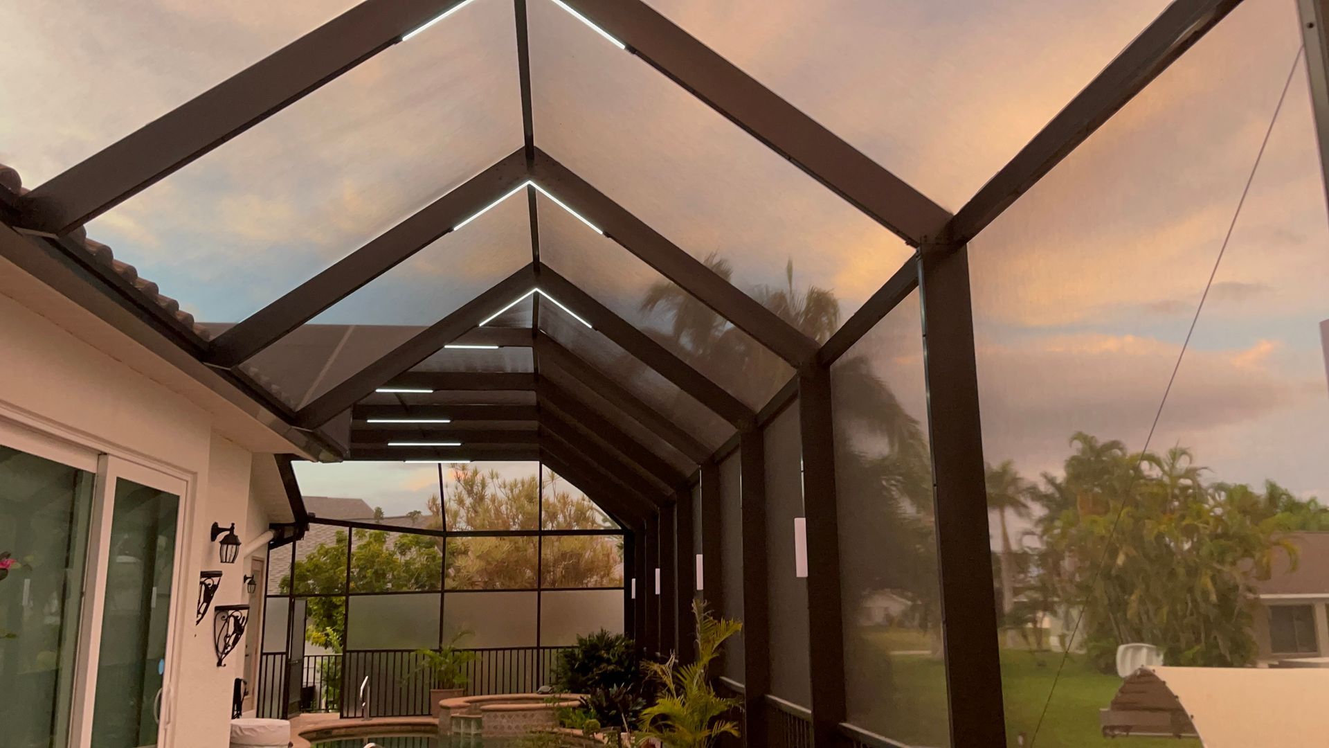Screened-in patio with dark brown frame and clear roof panels, looking out at a cloudy sky and yard.