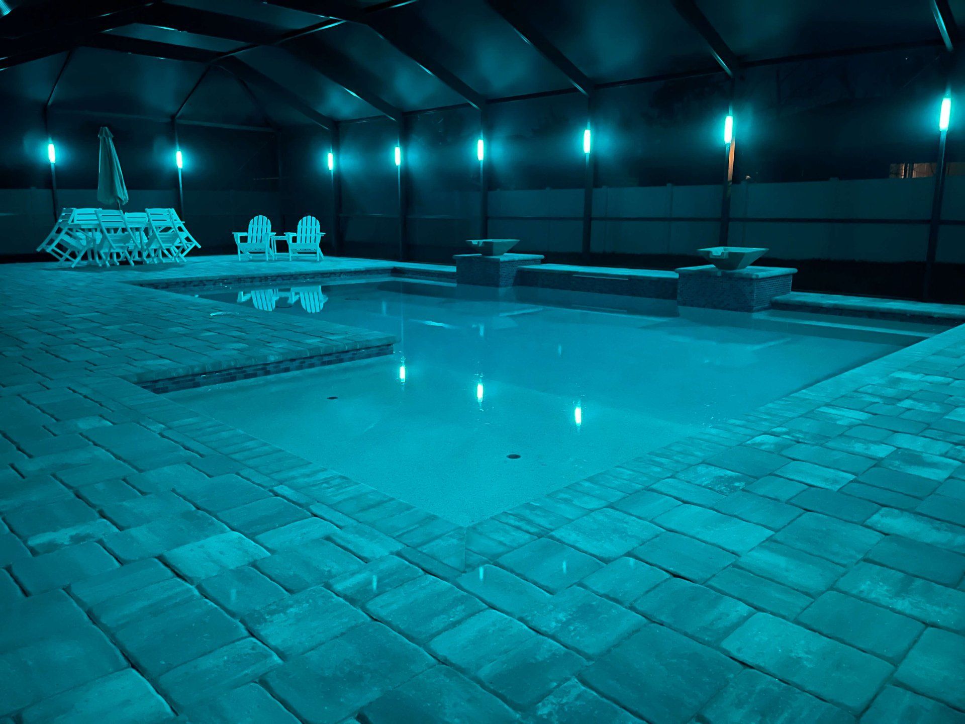 Indoor pool illuminated with blue lights, surrounded by brick pavers, chairs, and umbrella.