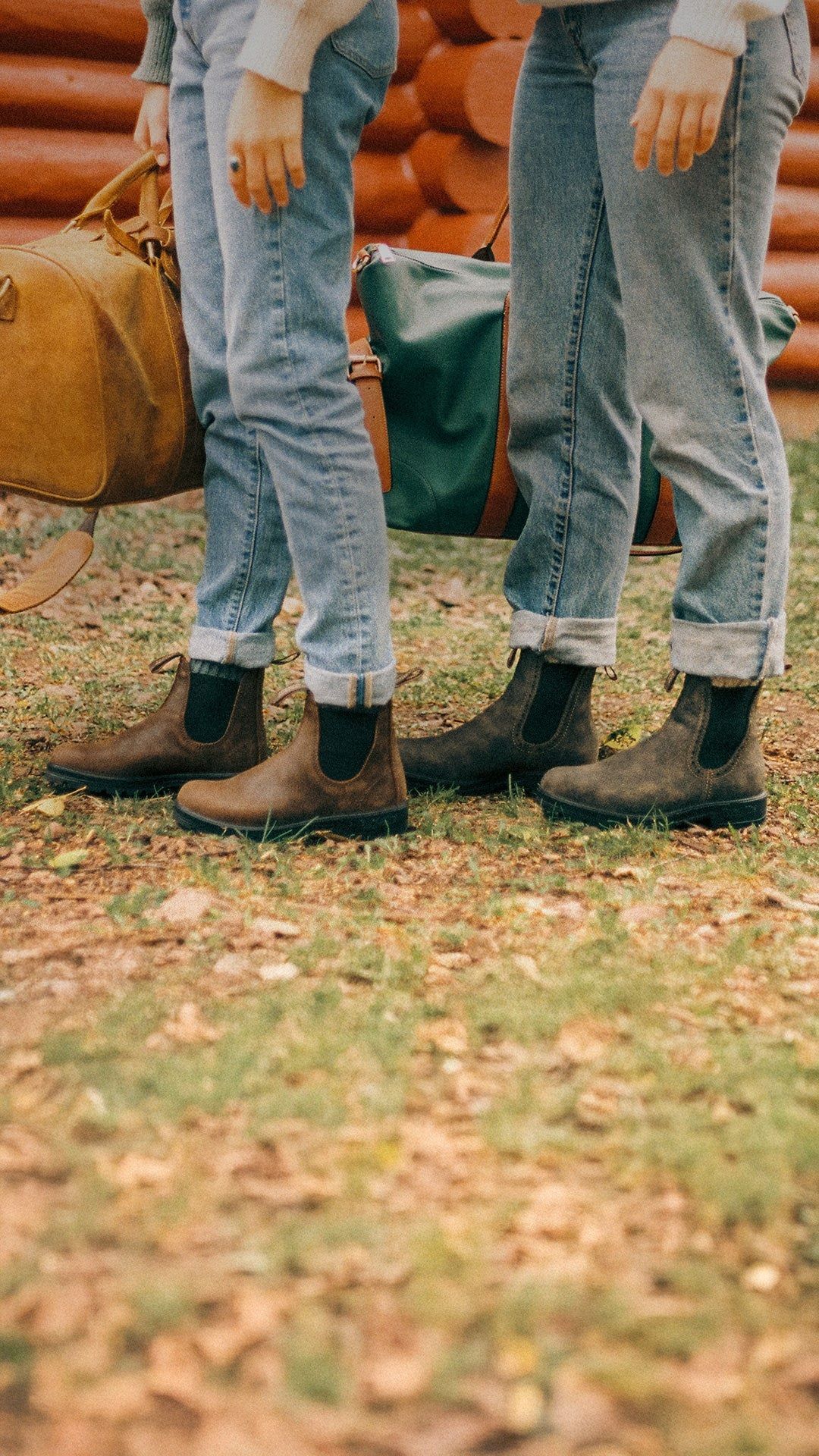 Two people wearing jeans and boots standing on grass, holding luggage in front of a log cabin.