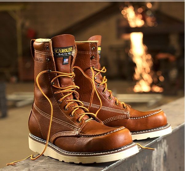 Brown leather work boots with white soles, yellow laces, and a blurred industrial background.