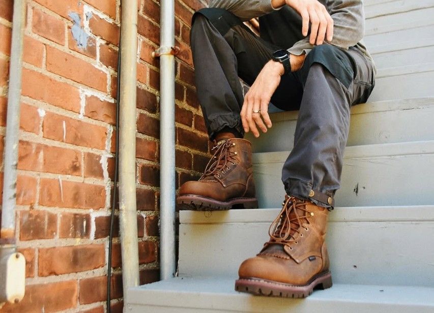 Person sitting on steps wearing work boots and gray pants. Brick wall background.