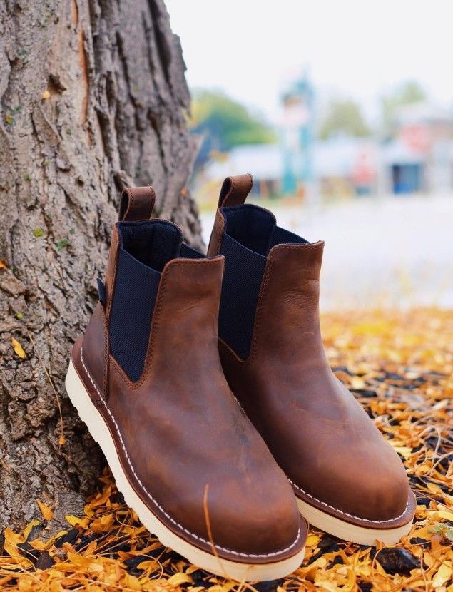 Brown leather Chelsea boots with white soles, leaning against a tree trunk in an outdoor setting.