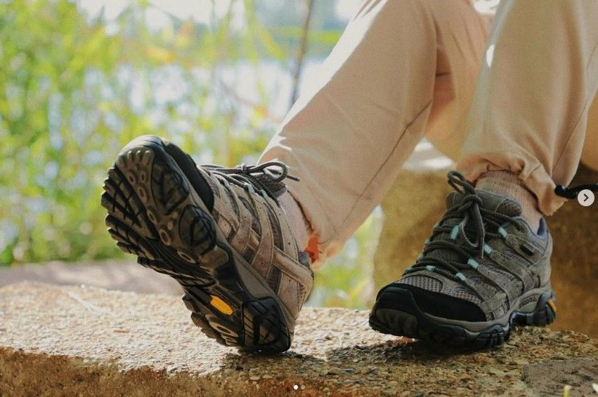 Person's feet in hiking boots resting on a stone ledge. Beige pants. Green background.