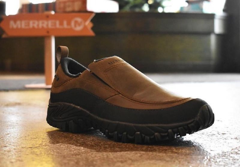 Brown Merrell slip-on shoe with a black sole, on a reflective surface, with a Merrell sign in the background.