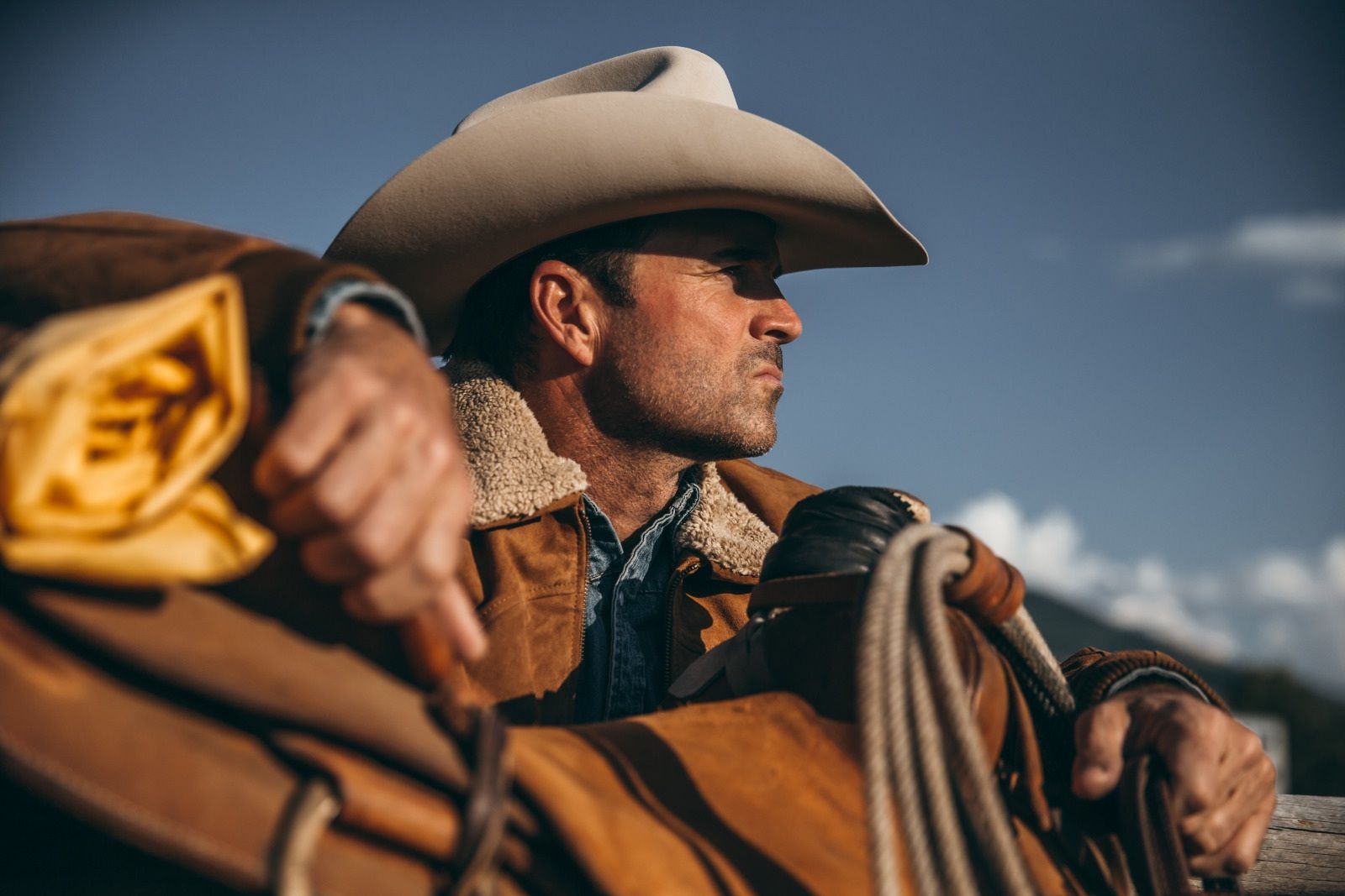 Cowboy wearing hat, jacket, looking away, holding saddle.