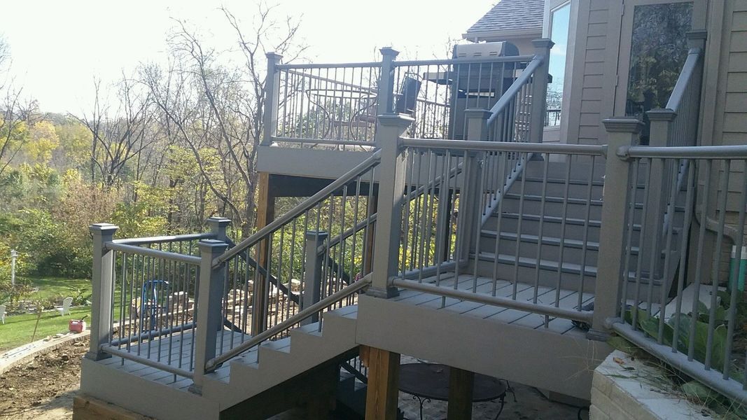 Gray deck with stairs and railings leading to a house with a wooded background.