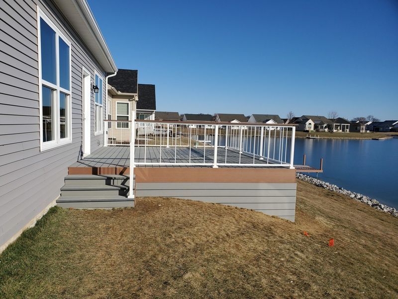 Deck on a house overlooking a lake, with gray siding, white railing, and brown base.