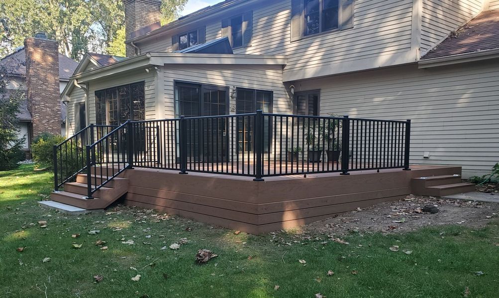 Backyard deck with black railing and steps attached to a light brown house with a sunroom.