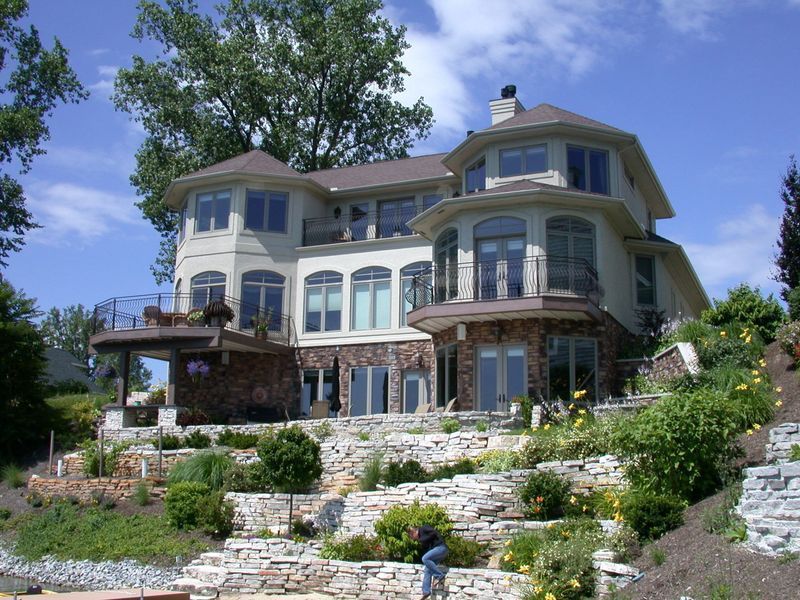 Large, multi-story beige house with stone accents, overlooking a landscaped hillside and water on a sunny day.