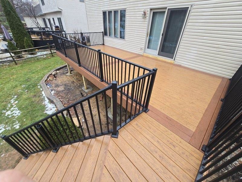 Wooden deck with black railings and steps leading to a house, surrounded by lawn and landscaping.