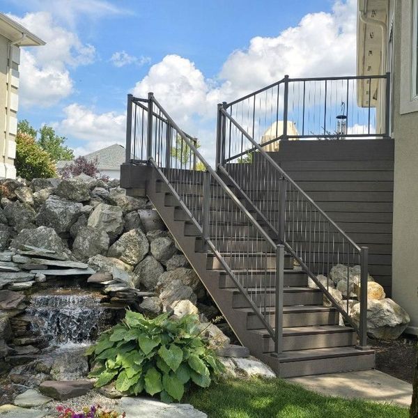 Staircase with metal railings leads to a deck. A rock waterfall is in the foreground, with blue sky.
