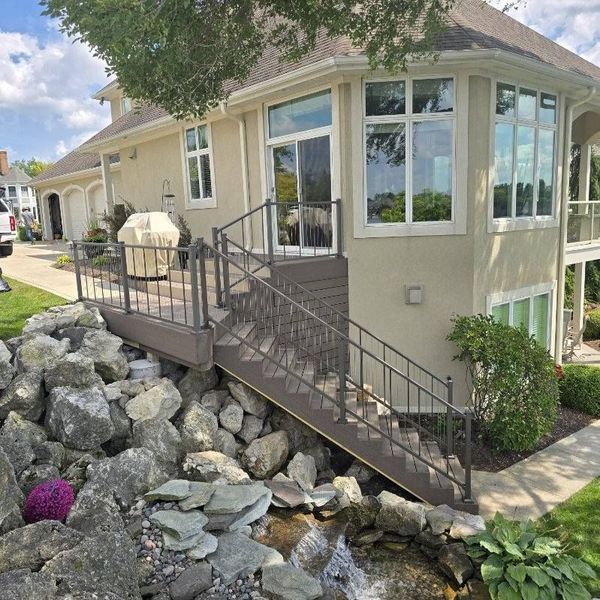 Deck with gray railings and stairs leads to a house with large windows. Rocks and a small waterfall in foreground.