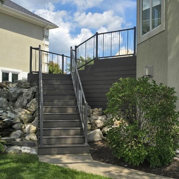 Exterior view of stairs leading up to a deck with black railing, beside a rock wall and bushes.