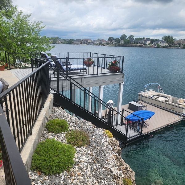 Deck overlooking a lake with boat dock. Black railings, gray decking, blue water, and cloudy sky.