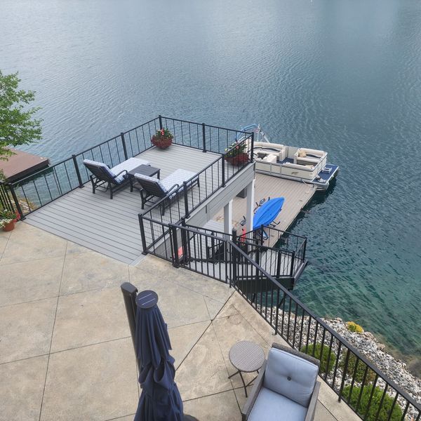 Deck overlooking a lake with boat dock. Gray deck, black railings, lounge chairs, and umbrella. Water visible.