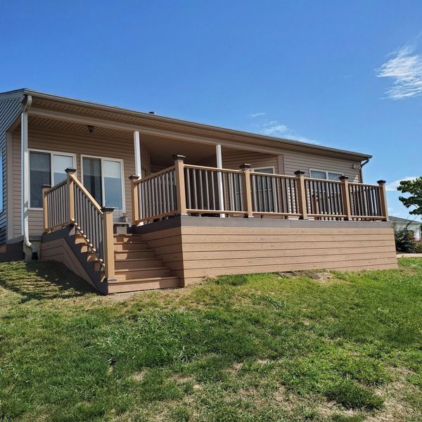 Tan house with a wooden deck and stairs, brown railings, and a green lawn under a blue sky.