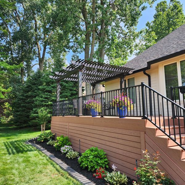 Brown deck with pergola, black railing, and flower pots, in a backyard.