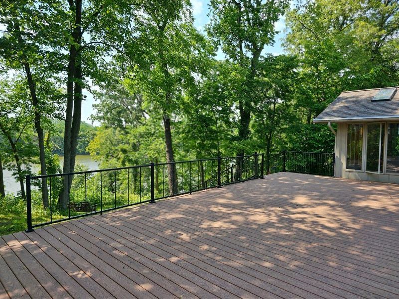 Wooden deck with black railing overlooking a lake, trees in the background, and a small structure on the right.