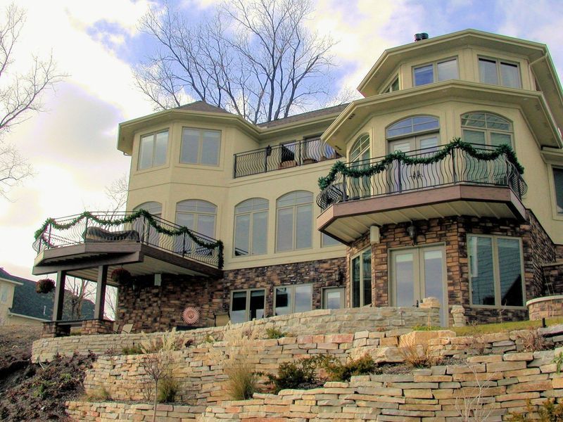 Two-story beige house with stone facade, two balconies with greenery, and a terraced landscape.