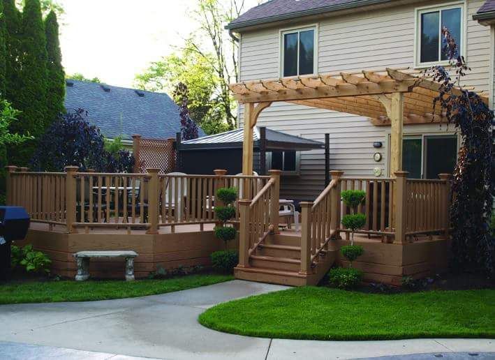 Wooden deck with pergola, stairs, and railing, beside a house with lawn and garden.